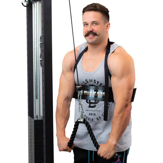 A man with a mustache, dressed in a gray tank top and black shorts, uses the Bells of Steel Arm Blaster for an intense arm workout. He holds the equipment with both hands, ready for biceps training against a white background.