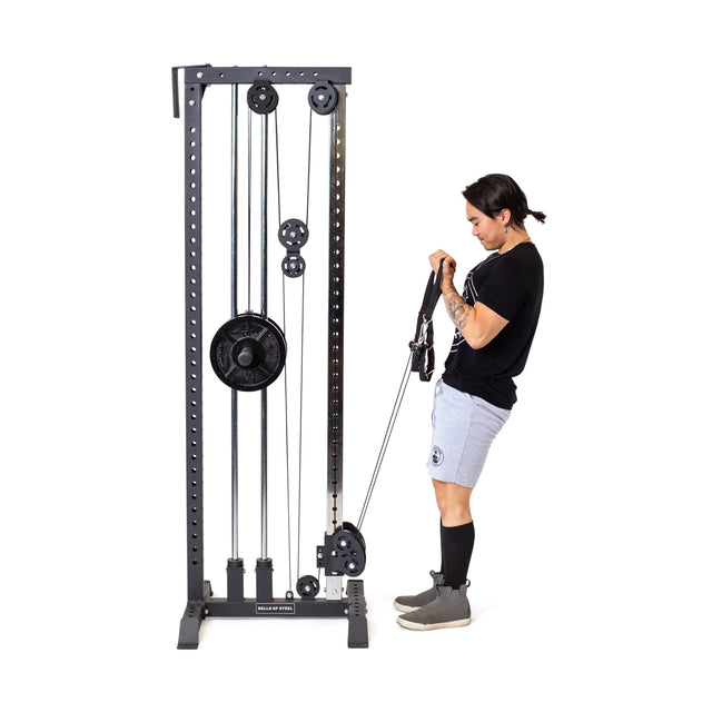 A person stands on a white background wearing a black t-shirt, gray shorts, and boots, using the Bells of Steel Cable Tower with adjustable handles for an arm workout.