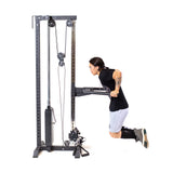 A person with long hair and tattoos performs bodyweight dips on parallel bars with adjustable handles, attached to the Bells of Steel Cable Tower, wearing a black shirt, gray shorts, and sneakers, against a white background.