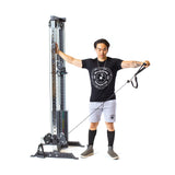 A person uses the Bells of Steel Cable Tower, gripping its adjustable handle with one arm outstretched to the side. They wear a black t-shirt, gray shorts, and black knee-high socks against a white background.