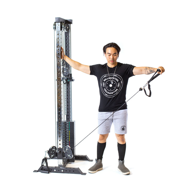 A person uses the Bells of Steel Cable Tower, gripping its adjustable handle with one arm outstretched to the side. They wear a black t-shirt, gray shorts, and black knee-high socks against a white background.