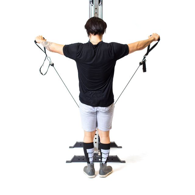 A person in a black shirt, gray shorts, and patterned knee-high socks uses the Bells of Steel Cable Tower for reverse fly exercises, facing away against a white background.