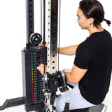 A person in a black shirt and gray shorts adjusts the weight stack on a Bells of Steel Cable Tower at the gym, selecting color-coded weight plates labeled in pounds for their workout.