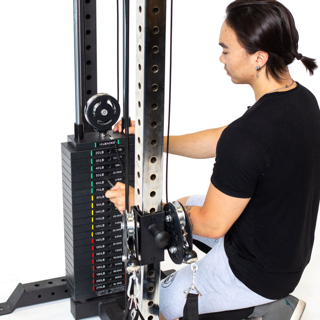 A person in a black shirt and gray shorts adjusts the weight stack on a Bells of Steel Cable Tower at the gym, selecting color-coded weight plates labeled in pounds for their workout.