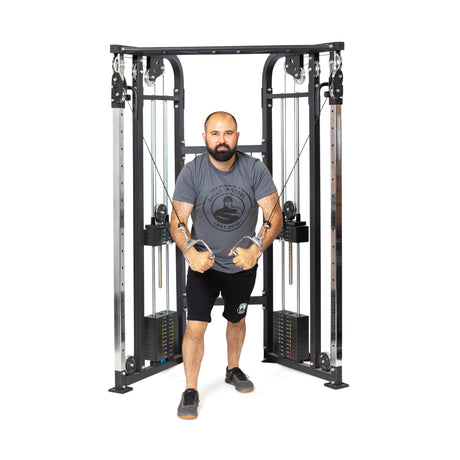 A bearded man exercises with focus on a Bells of Steel Functional Trainer. In a gray T-shirt and black shorts, he skillfully pulls the cables to his chest against a plain white background, highlighting the sleek design of the weight stacks and equipment.
