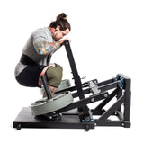 A tattooed person in a gray shirt, black shorts, knee sleeves, and lifting belt showcases impressive lower body strength on a Belt Squat Machine by Bells of Steel in a minimalist home gym with a clean white background.