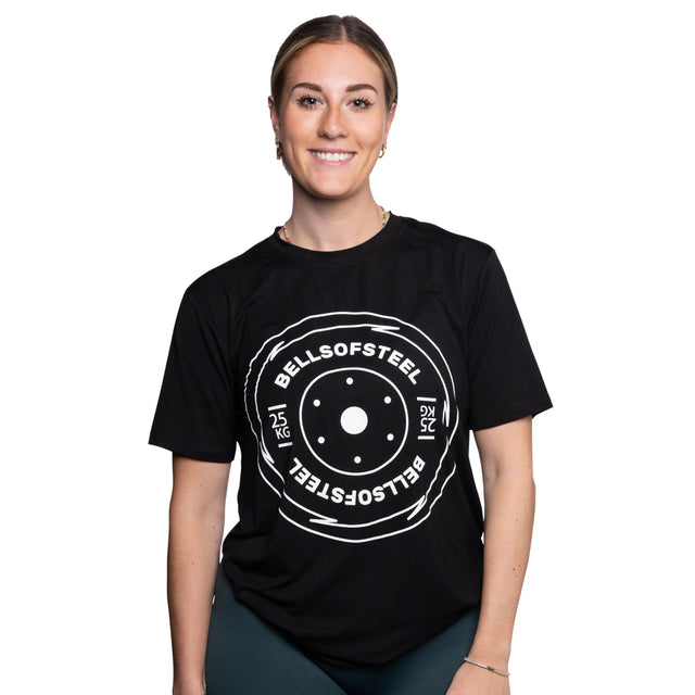 A woman smiles in a Bells of Steel Bamboo T-Shirt, which features a white "BELLS OF STEEL 25 KG" weight plate graphic on the front. She stands against a plain white background.