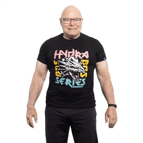 An older, bald man with glasses smiles against a white background. He sports a black Bells of Steel Bamboo T-Shirt featuring colorful "Hydra Bass Series" text encircling an illustrated dragon head.