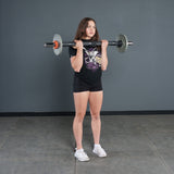 A woman in a black t-shirt and shorts stands indoors on a gray floor, holding a Bells of Steel Fat Bar - Olympic Curl Bar Cable Attachment at her shoulders as if preparing for a front squat or overhead press.