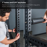 A man adjusts a bolt on the Bells of Steel Manticore Flat Foot Power Rack - Prebuilt (3" x 3", 1" Holes), while a woman holds a padded attachment near the rack, ideal for any home gym. Text above describes the keyhole sizes and compatibility for this versatile 3x3 power rack.