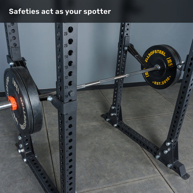 A loaded barbell rests on a squat rack inside a gym, with safety bars in place. Text at the top reads, "Safeties act as your spotter." The Bells of Steel Manticore Flat Foot Power Rack – Prebuilt (3" x 3", 1" Holes) is the perfect addition to any home gym, delivering stability and reliability for your heaviest lifts.