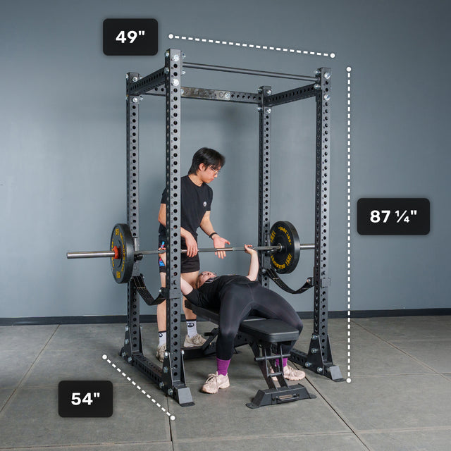 Two people in a gym use the Bells of Steel Manticore Flat Foot Power Rack - Prebuilt (3" x 3", 1" Holes) for bench pressing—one lifting while the other spots. Dotted lines outline the power rack’s dimensions: 49" wide, 54" deep, and 87¼" tall against a gray background, making it an ideal addition to your home gym setup.