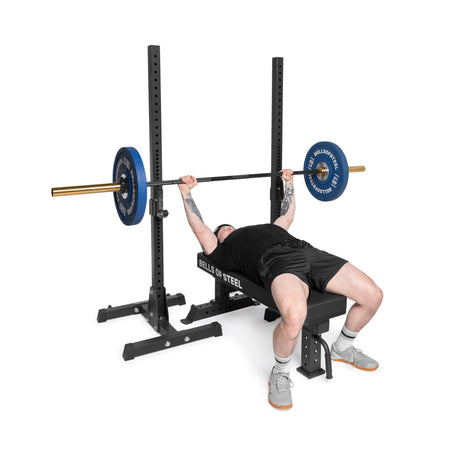 A person in a black sleeveless shirt, black shorts, and gray sneakers performs a bench press on the durable Bells of Steel Powerlifting Flat Bench using vibrant blue weights with a branded logo, showcasing an impressive 1,000lb max capacity.