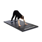 A person in black clothing stretches on a Bells of Steel Four Fold Mat, with hands and feet on the mat and hips raised against a plain white background.