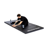 A person in black workout clothes stretches on a large, non-slip Four Fold Mat from Bells of Steel, set against a white background.