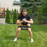 A woman exercises outdoors on the grass, sitting on a bench and wearing a black Bells of Steel Glute Loop resistance band positioned above her knees, with her hands clasped in front of her chest. She sports a black shirt, shorts, and white sneakers—ideal for an effective booty workout.