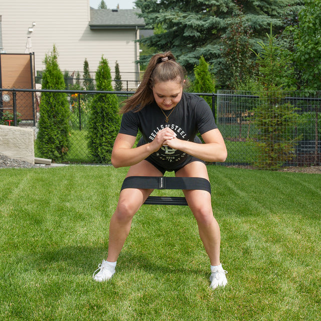 A woman exercises outdoors on the grass, sitting on a bench and wearing a black Bells of Steel Glute Loop resistance band positioned above her knees, with her hands clasped in front of her chest. She sports a black shirt, shorts, and white sneakers—ideal for an effective booty workout.