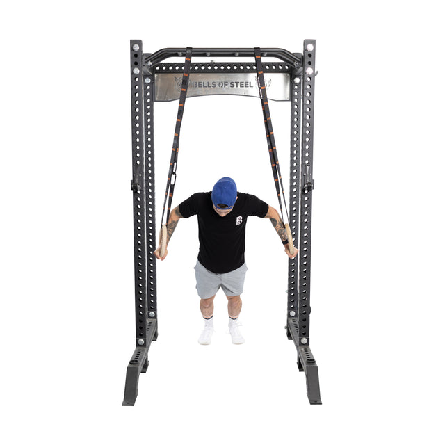 A person in a black T-shirt, gray shorts, and a blue cap is immersed in bodyweight training using Bells of Steel Gymnastics Rings attached to a sturdy metal rack. Positioned in a plank-like stance, they're executing an intensive core workout.