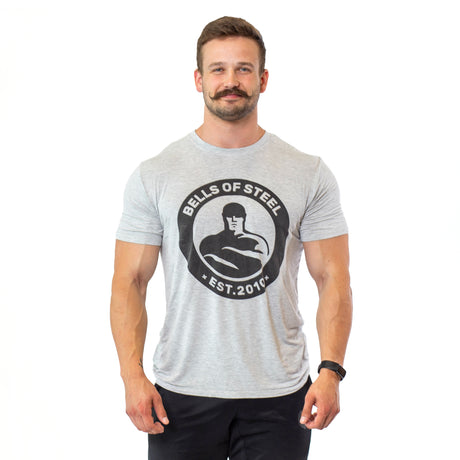 A smiling man with a mustache faces forward in black pants and a light gray, sweat-wicking Bells of Steel Bamboo T-Shirt, standing against a white background.