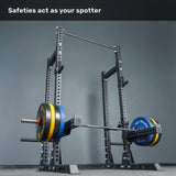 A loaded barbell sits securely on the safety arms of the Bells of Steel Manticore Half Rack Builder (3" x 3", 1" Holes) with robust uprights. The text above states, "Safeties act as your spotter." The plain gray wall backdrop highlights this ideal home gym setup.