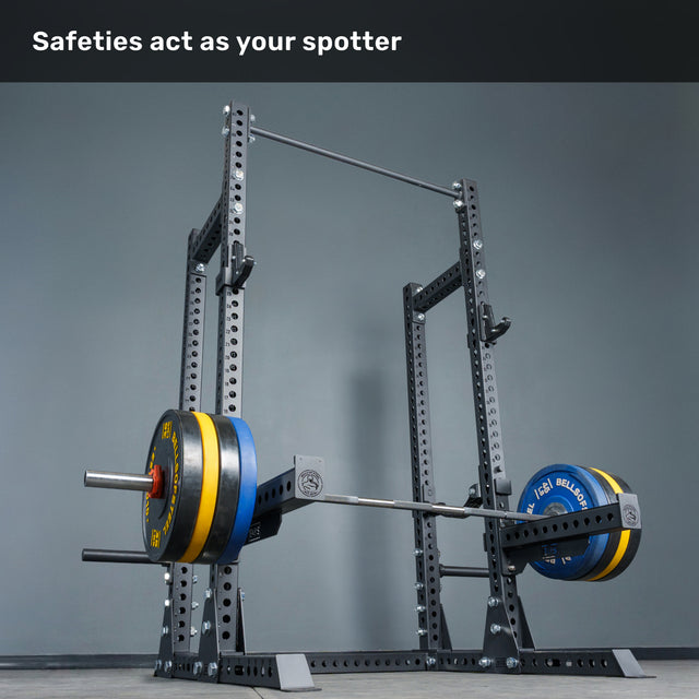 A loaded barbell sits securely on the safety arms of the Bells of Steel Manticore Half Rack Builder (3" x 3", 1" Holes) with robust uprights. The text above states, "Safeties act as your spotter." The plain gray wall backdrop highlights this ideal home gym setup.