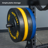 Close-up of weight plates in black, blue, and yellow neatly stacked on a storage rack, set against the gray walls and floor of a gym environment. The scene highlights the ideal setup for a home gym or the Bells of Steel Manticore Half Rack - Prebuilt (3" x 3", 1" Holes) arrangement.