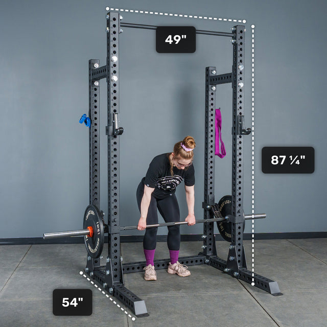 A person prepares to lift a barbell inside the Bells of Steel Manticore Half Rack - Prebuilt (3" x 3", 1" Holes), which features robust 3x3 uprights. The rack’s dimensions are clearly labeled: 49 inches wide, 54 inches deep, and standing at 87 ¼ inches tall—making it an ideal choice for any serious home gym setup. Purple and blue bands hang on the rack.