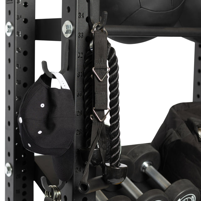 A close-up of the Bells of Steel Utility Hook on a gym rack, storing a black baseball cap, a black tricep rope attachment, weights, and a medicine ball for efficient gym organization.