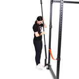 A woman in athletic wear adjusts a barbell on a Bells of Steel Vertical Mount Barbell Holder Rack Attachment, which is fixed to a black power rack with an orange safety arm, all on a white background.