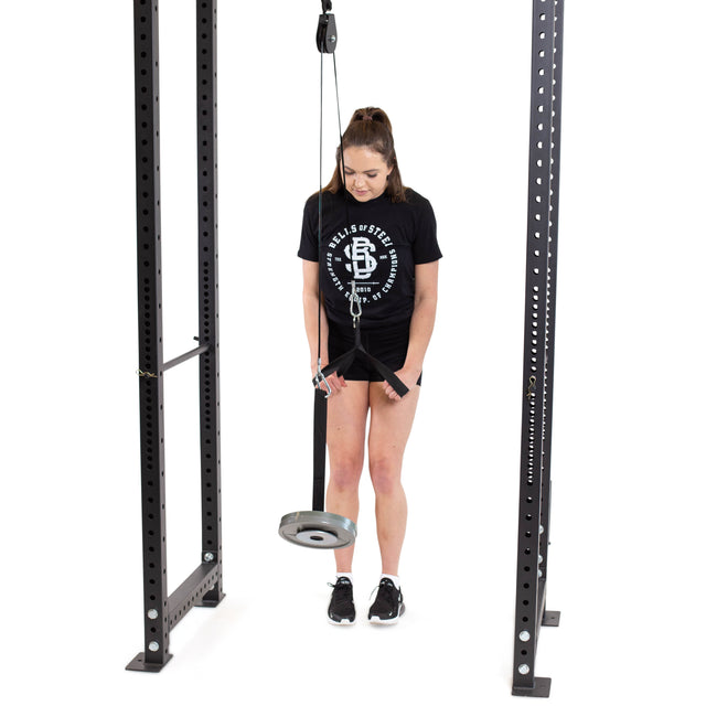 A woman in a black t-shirt, shorts, and athletic shoes stands in a squat rack, using the Bells of Steel Cable Pulley and wrist roller as she prepares for a forearm workout.