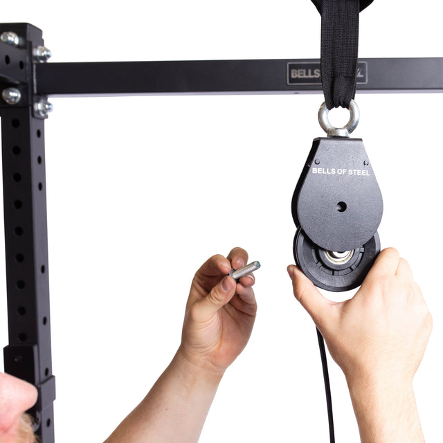 A person installs a Bells of Steel Cable Pulley onto a black metal gym rack, holding a bolt or pin in one hand and the Cable Pulley in the other—ideal for lat pulldowns and tricep pushdowns.