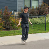 Wearing black athletic clothes, a person does cardio training with the Bells of Steel Jump Rope on a sidewalk in front of a house with a fenced yard and greenery.