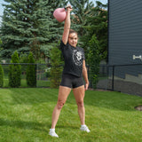 A person stands on grass, lifting a pink Bells of Steel Competition Kettlebell overhead with one arm. They are dressed in a black T-shirt, black shorts, and white shoes. In the background, there are trees, a dark house, and a wire fence.