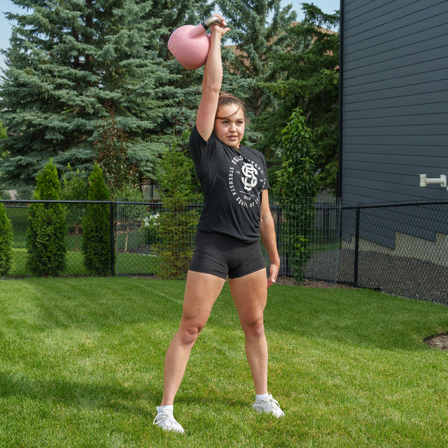 A person stands on grass, lifting a pink Bells of Steel Competition Kettlebell overhead with one arm. They are dressed in a black T-shirt, black shorts, and white shoes. In the background, there are trees, a dark house, and a wire fence.