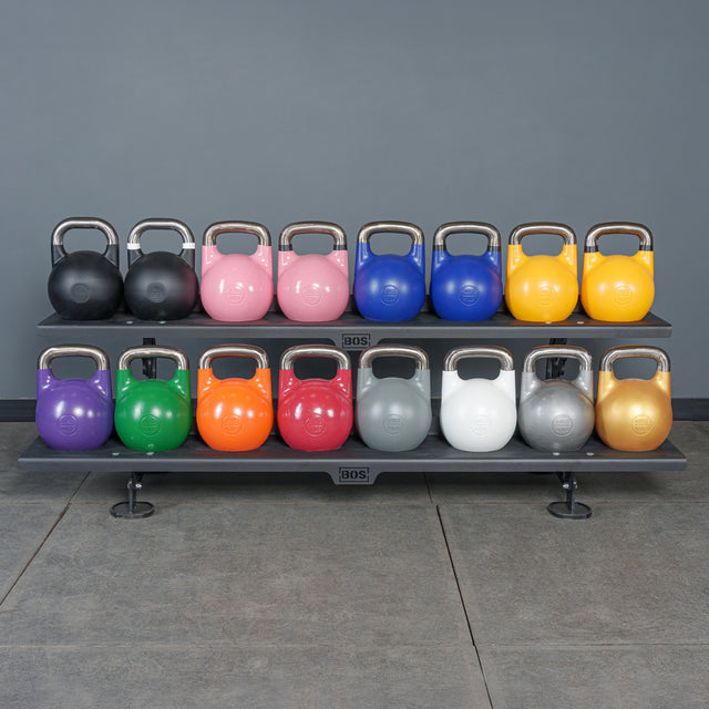 Two rows of Bells of Steel Competition Kettlebells, each color-coded by weight and size, are neatly arranged on a black rack against a gray wall—making it simple to select the right kettlebell to match any weight guide.