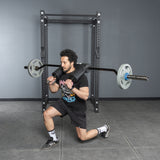 A man in athletic wear uses the Bells of Steel Safety Squat Bar – The SS4 for lunges, holding it across his shoulders in front of a squat rack, with his right knee bent forward and left leg extended behind.
