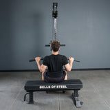 A person sits on a Bells of Steel bench, gripping the Fat Bar - Lat Pulldown Bar attached to a lat pulldown machine, ready to boost grip strength in a gym with gray walls and flooring.