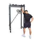A person dressed in a black shirt and shorts works out using a landmine attachment on the Bells of Steel Manticore Four Post Power Rack - Prebuilt (3" x 3", 1" Holes). They are positioned to press a barbell with a weight plate, utilizing its versatile design that is ideal for home gyms.