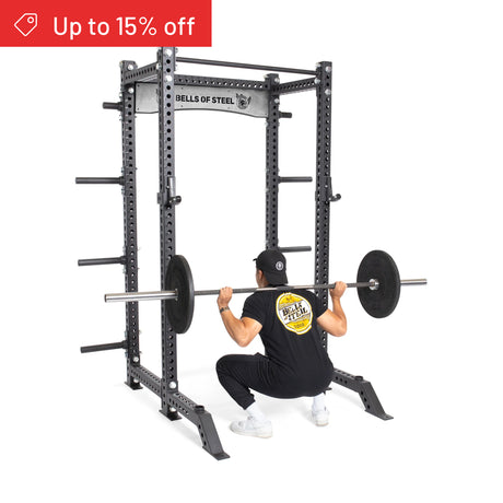 A person squats with a barbell inside the Bells of Steel Manticore Collegiate Power Rack Builder (3" x 3", 1" Holes), wearing a black shirt and cap. A red "Up to 15% off" banner appears—ideal for any modular home gym.