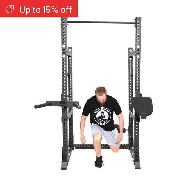 A man in a black t-shirt and gray shorts does lunges inside a Bells of Steel Manticore Half Rack Builder (3" x 3", 1" holes). A red banner in the top left corner says "Up to 15% off.