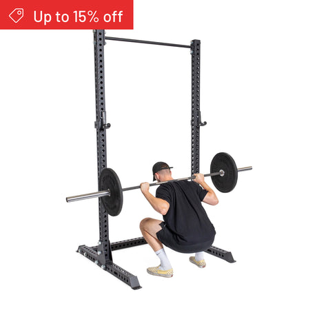 A person in athletic wear performs a barbell squat on the Bells of Steel Manticore Squat Stand Builder (3" x 3", 1" Holes). A red banner says "Up to 15% off." The white background is perfect for home gym inspiration.