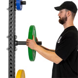 A man in a black shirt and cap, sporting a beard, is expertly placing a green Olympic-sized weight plate onto a black power rack featuring numbered indicators. He adjusts the Bells of Steel Bolt-On Plate Pegs for balance alongside other plates.
