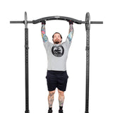 In a gym, a tattooed person in a gray shirt and black shorts performs a pull-up on a squat rack equipped with the Bells of Steel Arch Nemesis Swiss Bar.