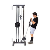 Wearing athletic gear, a person uses the Bells of Steel Cable Tower, adjusting handles to pull cables connected to a weight stack while exercising indoors against a white background.
