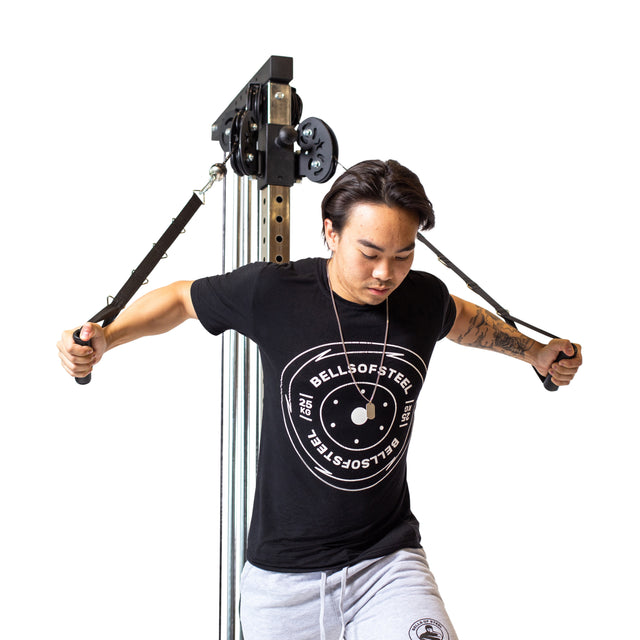 A person wearing a black Bells of Steel t-shirt demonstrates a chest fly using the Bells of Steel Cable Tower, standing with one foot forward and gripping the adjustable handles. The background is plain white.