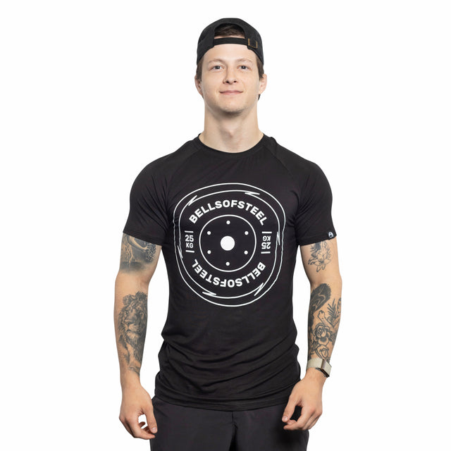 A man wearing a backward black cap and a Bells of Steel Bamboo T-Shirt with a weight plate graphic faces forward, smiling, with tattooed arms visible against a white background.