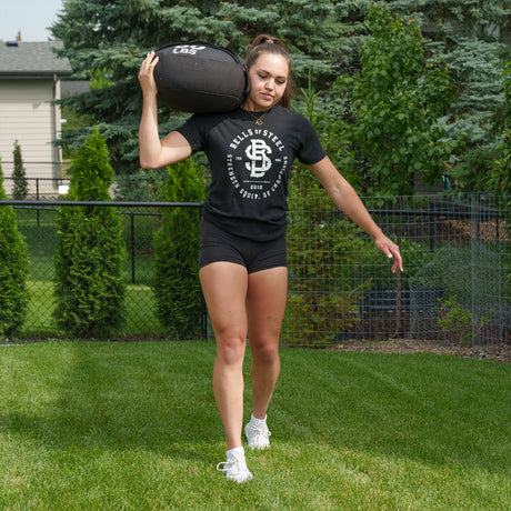 A woman in athletic clothing carries a heavy Bells of Steel Fitness Sandbag on her shoulder while walking on grass in a backyard with trees and bushes in the background.