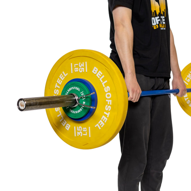 In black pants and shirt, a person holds a polished chrome barbell with large yellow "BELLS OF STEEL" 35 lb plates secured by Olympic barbell sleeves and grips the product from "Bells of Steel.