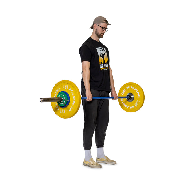 Wearing a black T-shirt, dark pants, and a cap, a man lifts a polished chrome barbell with yellow weights. He uses Bells of Steel's Spring Collars to keep the weights secure. Standing on a white background, he slightly faces left with his head down and arms fully extended.