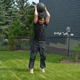Dressed in black workout gear, an athlete lifts a Bells of Steel Slam Ball overhead while standing on backyard grass, preparing to slam it down—a dynamic way to unleash explosive power and ramp up the intensity of your cardio workouts.